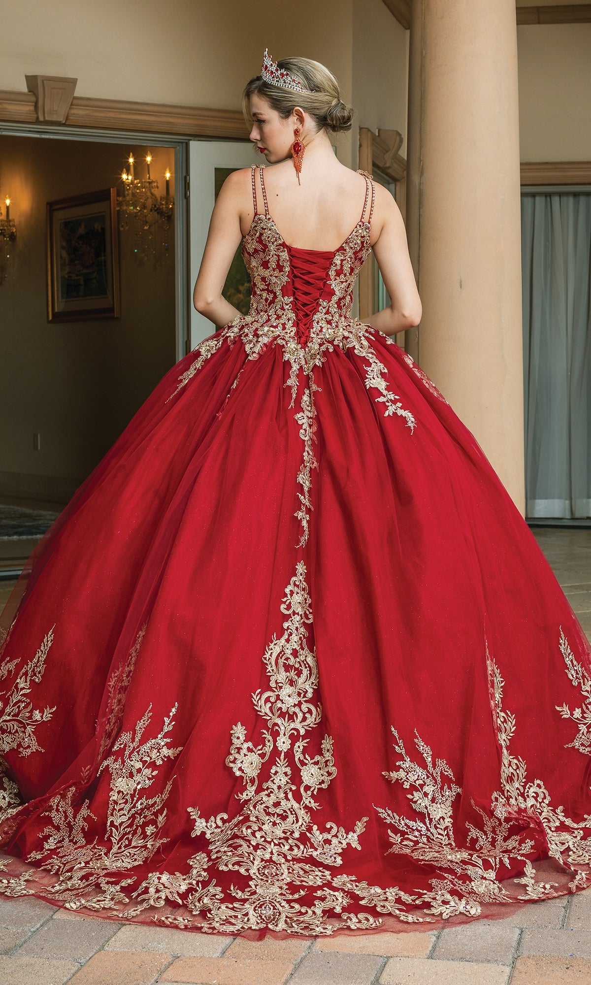 A woman in the Quinceanera Dress 1612 By Dancing Queen, a burgundy ball gown with gold lace embroidery, formal earrings, and a tiara stands outdoors near columns, facing away from the camera.